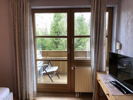 Una habitación con puerta de vidrio corredera que da a un balcón con una silla y una mesa. Hay un radiador en el suelo y un televisor sobre un mueble.