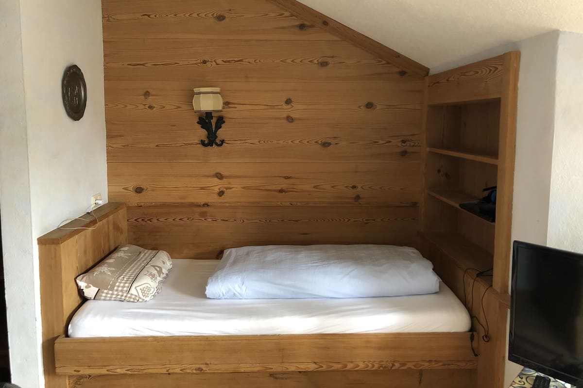 A wooden bunk bed with a white mattress and pillows, a wall-mounted lamp, and a television on a wooden shelf.