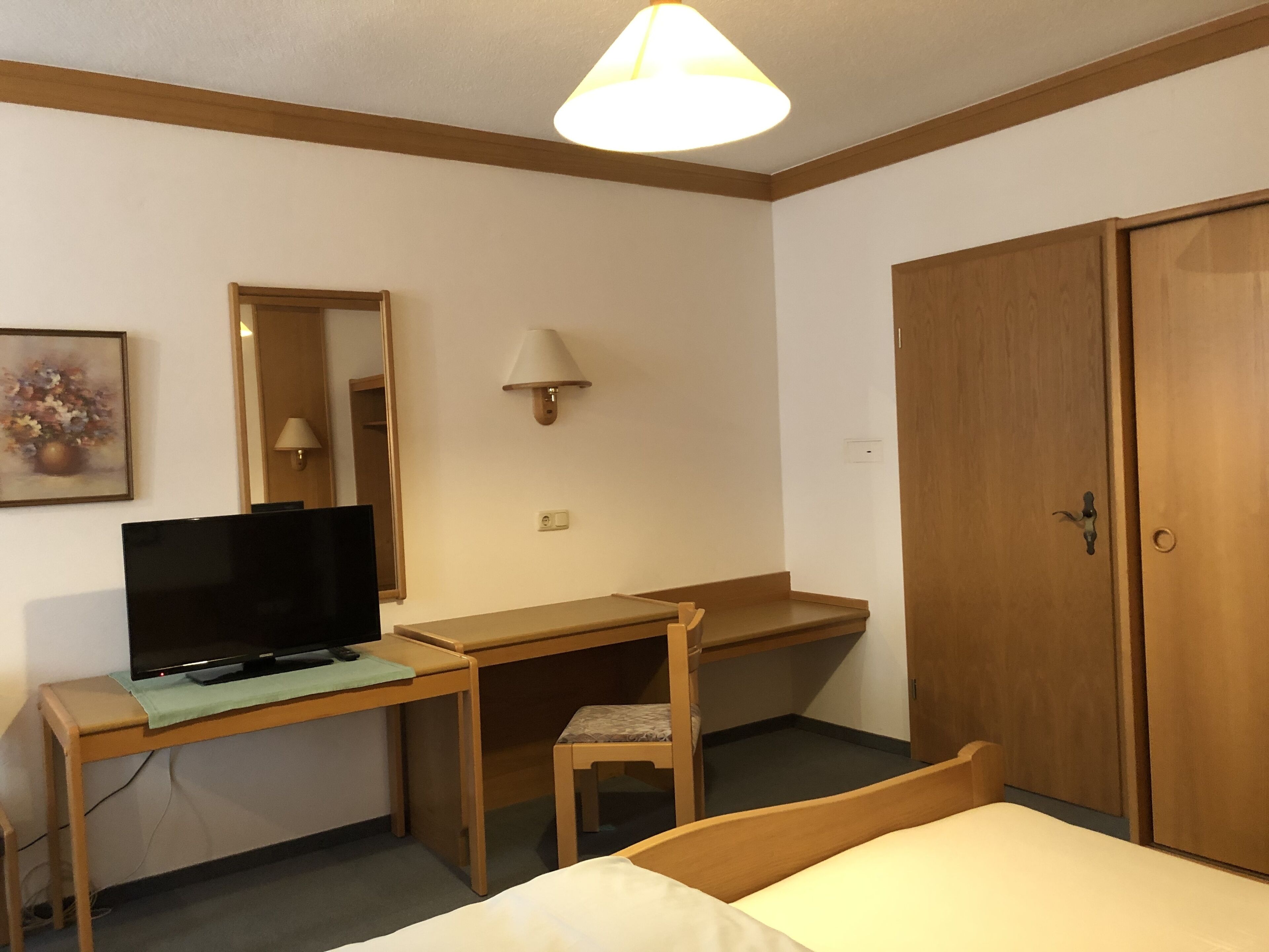 Quarto de hotel com cama, escrivaninha com computador, cadeira, televisão, luminária e guarda-roupa.
