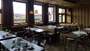 Un comedor con varias mesas y sillas, ventanas amplias con cortinas y lámparas colgantes.