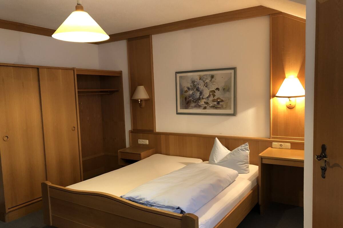 A hotel room with a wooden bed, a nightstand, a wall-mounted lamp, a framed picture, and a wardrobe.