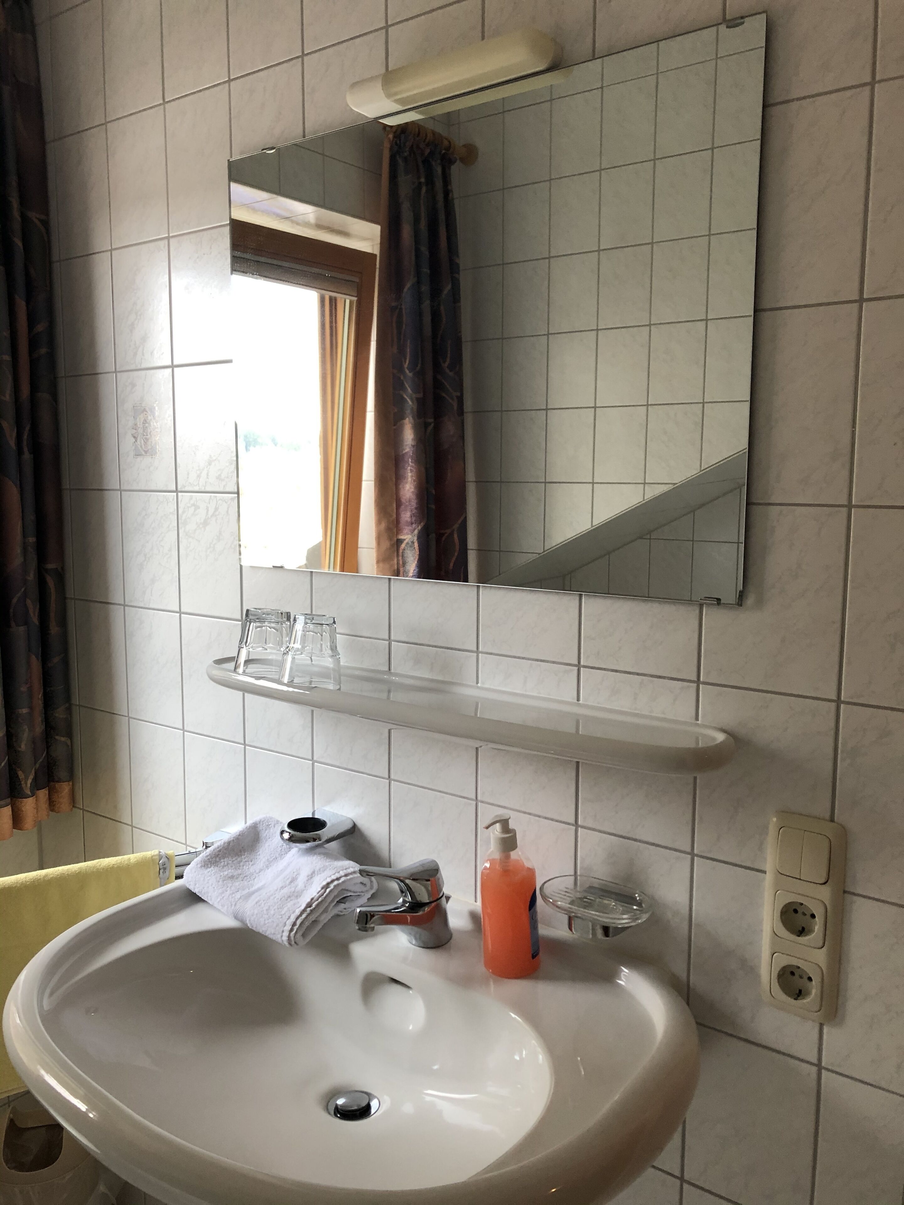 A bathroom with a white sink, a mirror above it, a towel hanging on the wall, and a shelf with glasses and a bottle.
