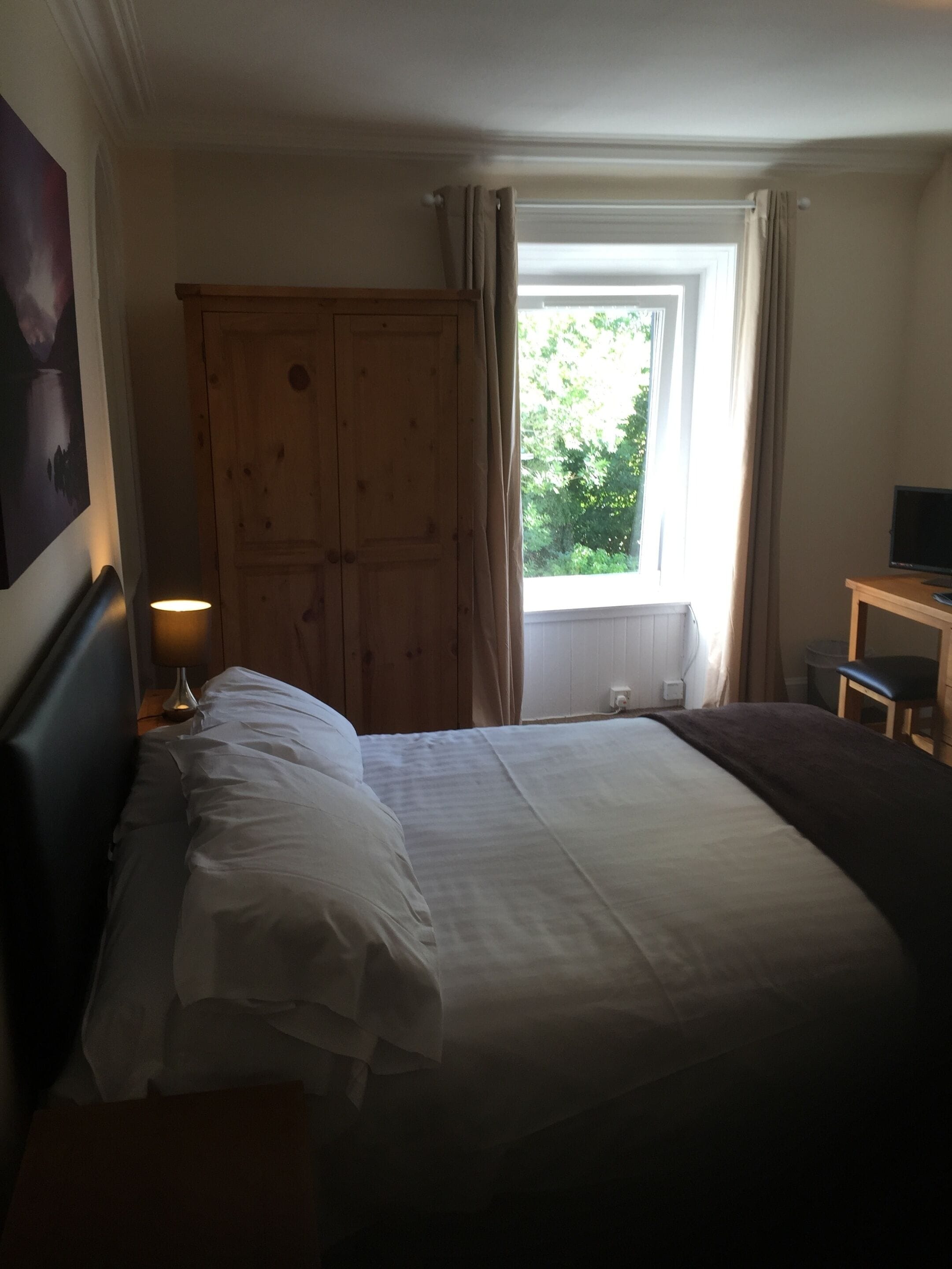 A hotel room with a large bed, a wooden wardrobe, a desk with a chair, and a window with curtains.