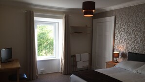 A hotel room with a bed, a desk with a TV, a window with curtains, a bedside lamp, and a patterned wallpaper. - The Shandwick Inn (Invergordon)