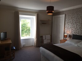 A hotel room with a bed, a desk with a TV, a window with curtains, a bedside lamp, and a patterned wallpaper.