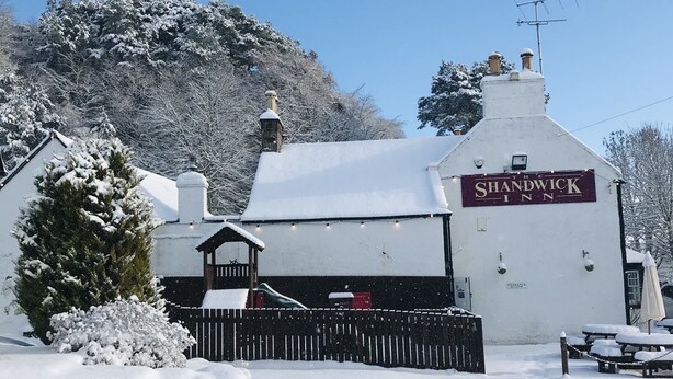 A snow-covered building labeled "SHANDWICK INN" with a stone wall entrance, a wooden fence, and a bench. - The Shandwick Inn (Invergordon)