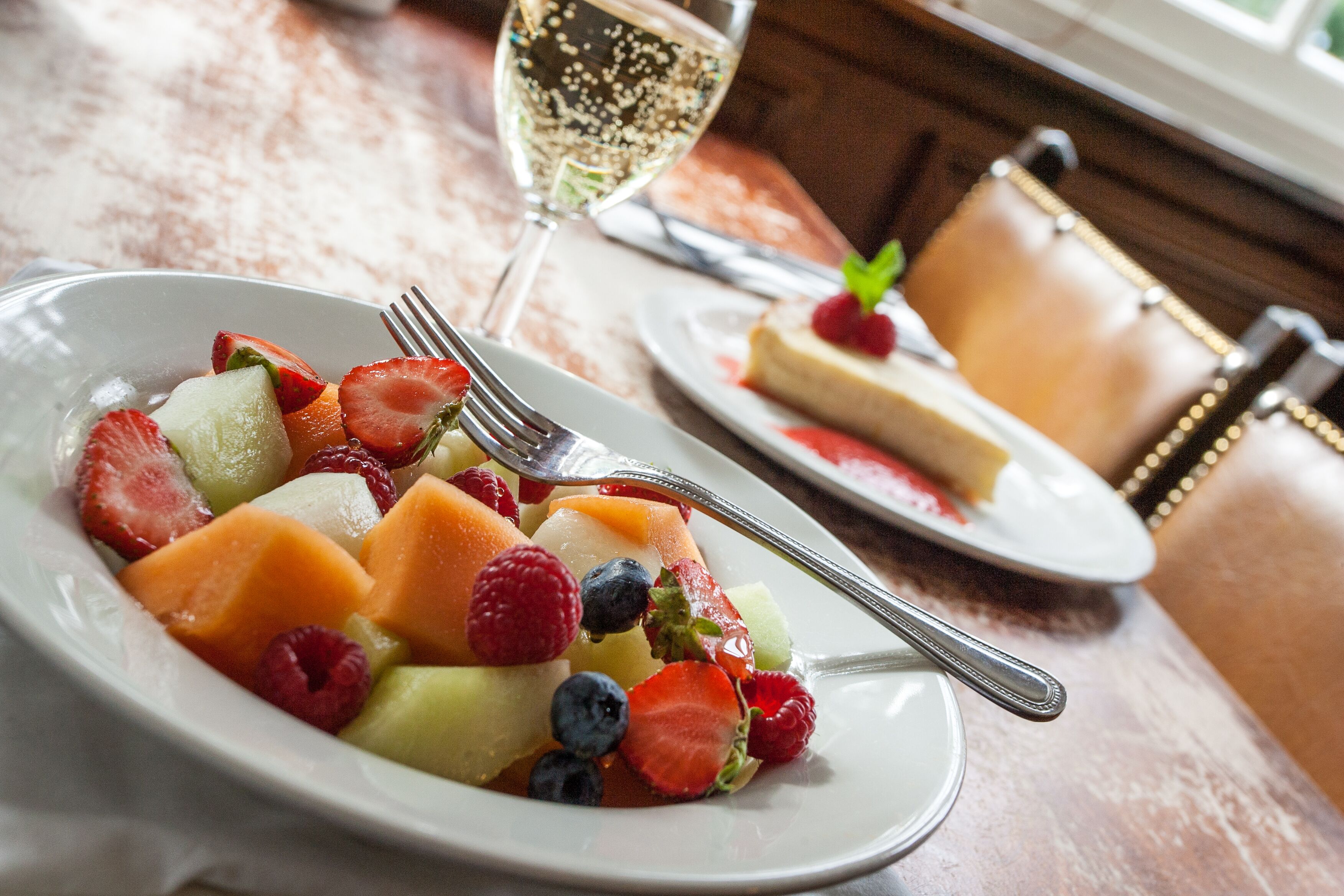 A plate of mixed fruit salad with a fork, a glass of white wine, and a slice of cheesecake on a table.