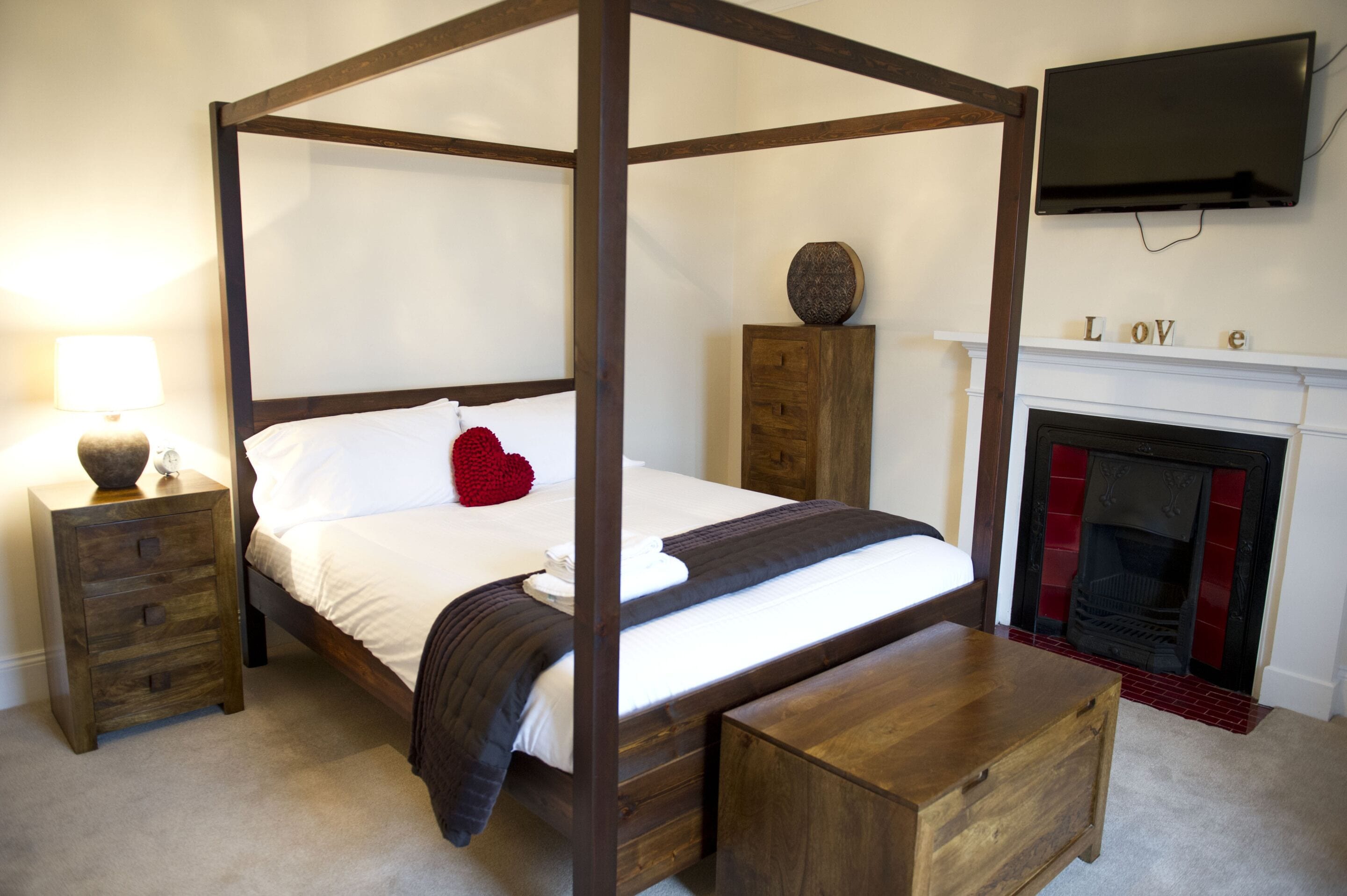 Una cama con dosel de madera, una mesita de noche con cajones, una lámpara de noche, un cojín con forma de corazón rojo, un pequeño mueble de madera y una chimenea con la palabra "LOVE" escrita en ella.