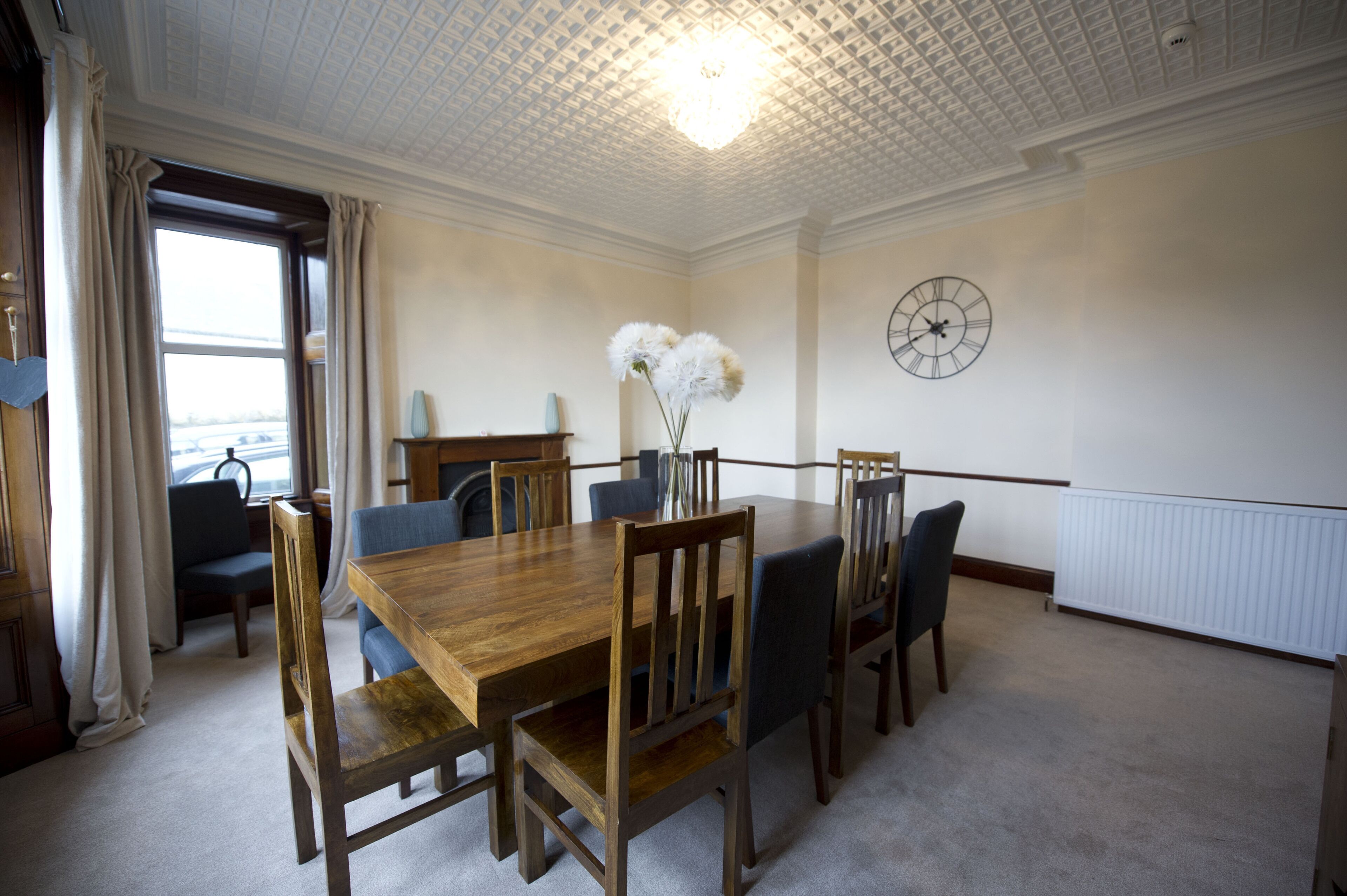 Een eetkamer met een houten tafel en stoelen, een vaas met witte bloemen, een klok aan de muur en een raam met gordijnen.