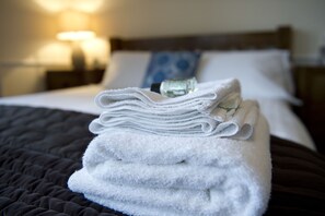 A neatly folded stack of white towels on a bed with a dark quilt, a bottle of lotion, and a blue pillow in a hotel room.