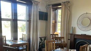 Uma área de refeições com mesas e cadeiras de madeira, janelas amplas com cortinas, uma lareira e uma televisão fixada na parede.