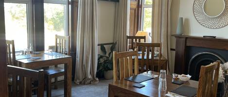 A dining area with wooden tables and chairs, large windows with curtains, a fireplace, and a television mounted on the wall.