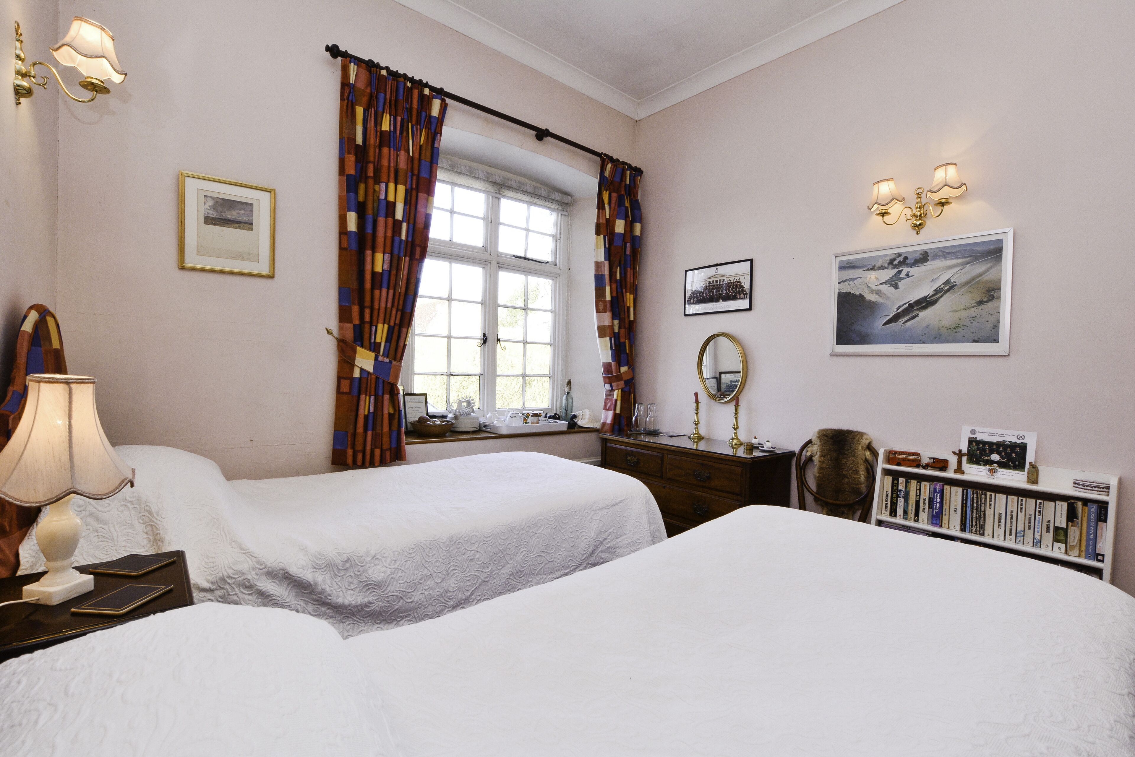A hotel room with two beds, a window with curtains, a bedside table with a lamp, a dresser, a chair, a bookshelf, and wall-mounted lights.