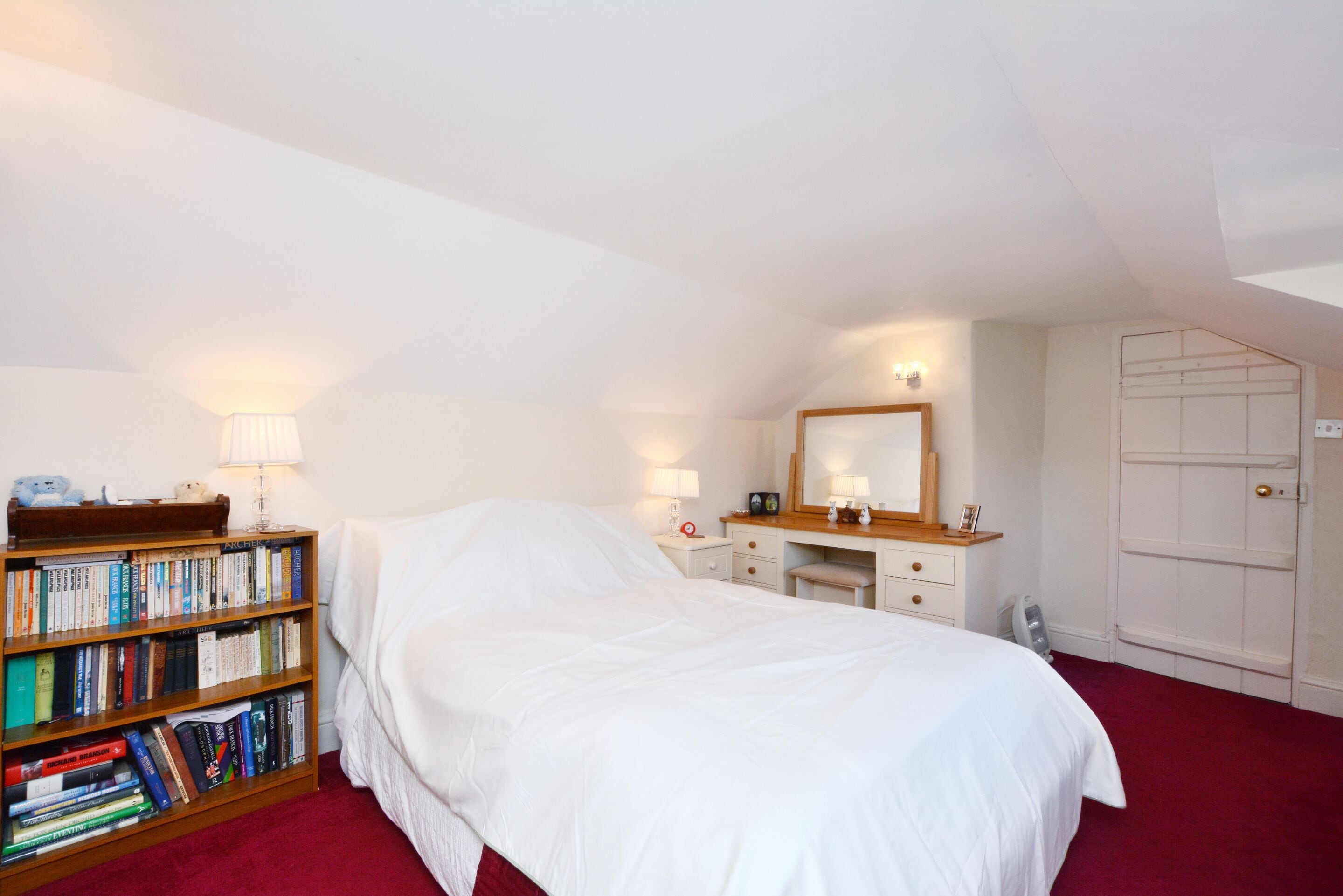 A bedroom with a large bed, a bedside table with a lamp, a dresser, a mirror, a wardrobe, and a bookshelf filled with books.