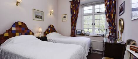 A hotel room with two beds, a window with curtains, a chair, a bookshelf, and wall-mounted lamps.