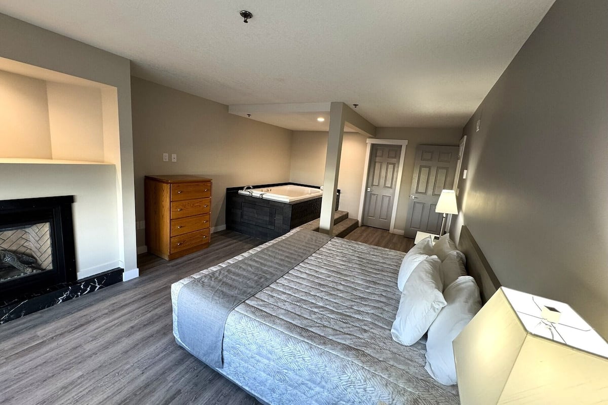 A hotel room with a large bed, a wooden dresser, a fireplace, a bathtub, and a door leading to another room.