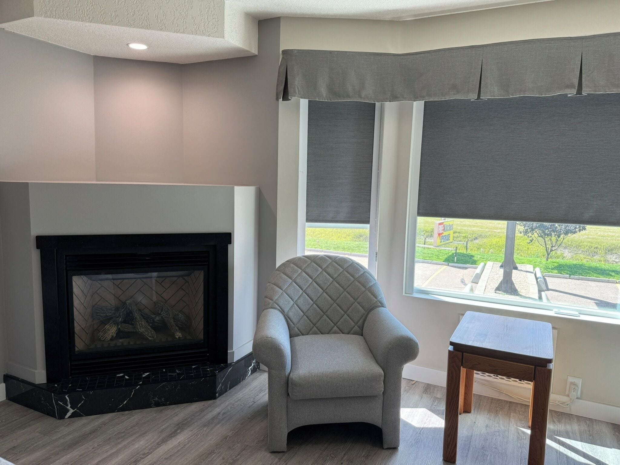 A living room with a fireplace, a comfortable armchair, a small wooden table, and a window with a view of a green landscape.