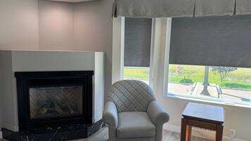 A living room with a fireplace, a comfortable armchair, a small wooden table, and a window with a view of a green landscape.