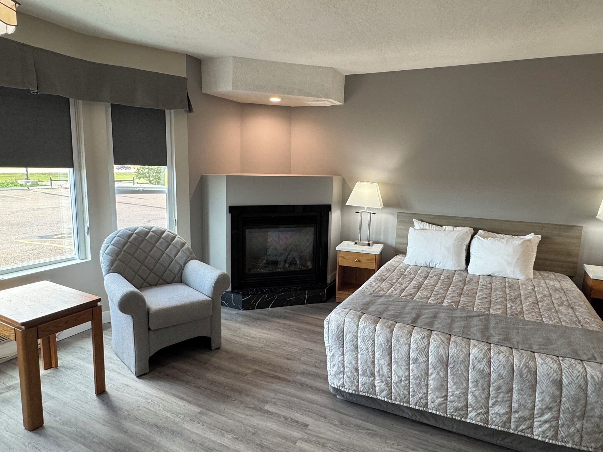 A hotel room with a bed, a chair, a small table, a nightstand, a lamp, and a fireplace.