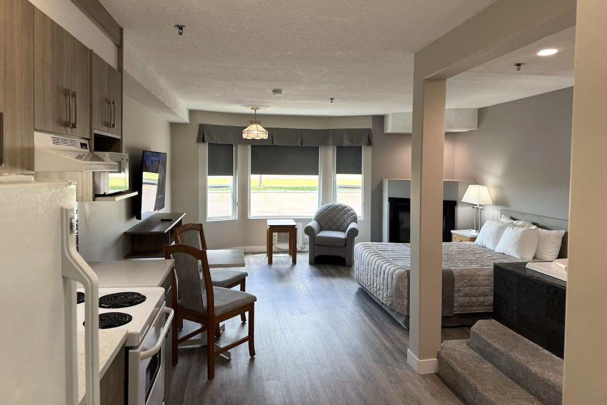 A compact hotel room with a kitchenette, a bed, a seating area with a chair and table, and a fireplace.