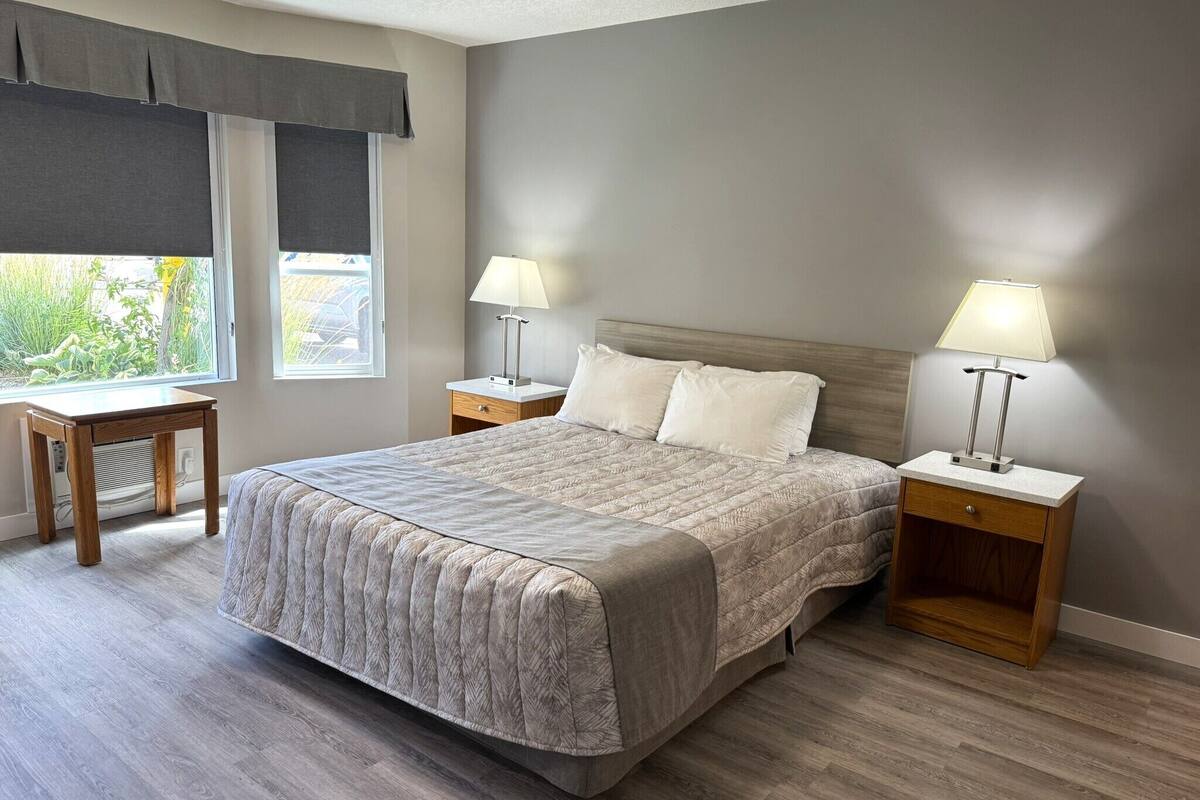 A bedroom with a large bed, two bedside tables with lamps, a small wooden nightstand, and a window with a view of greenery.