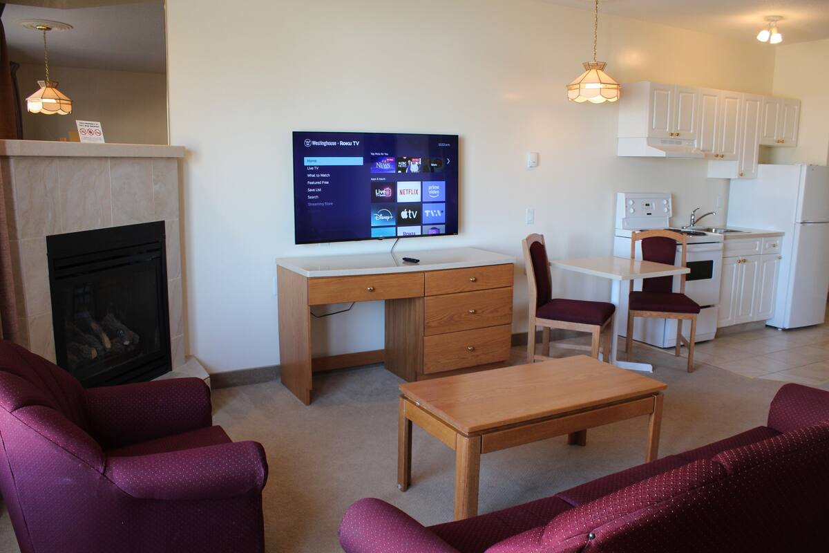 A living room with a fireplace, a TV mounted on the wall, a wooden desk, and a dining table with chairs.
