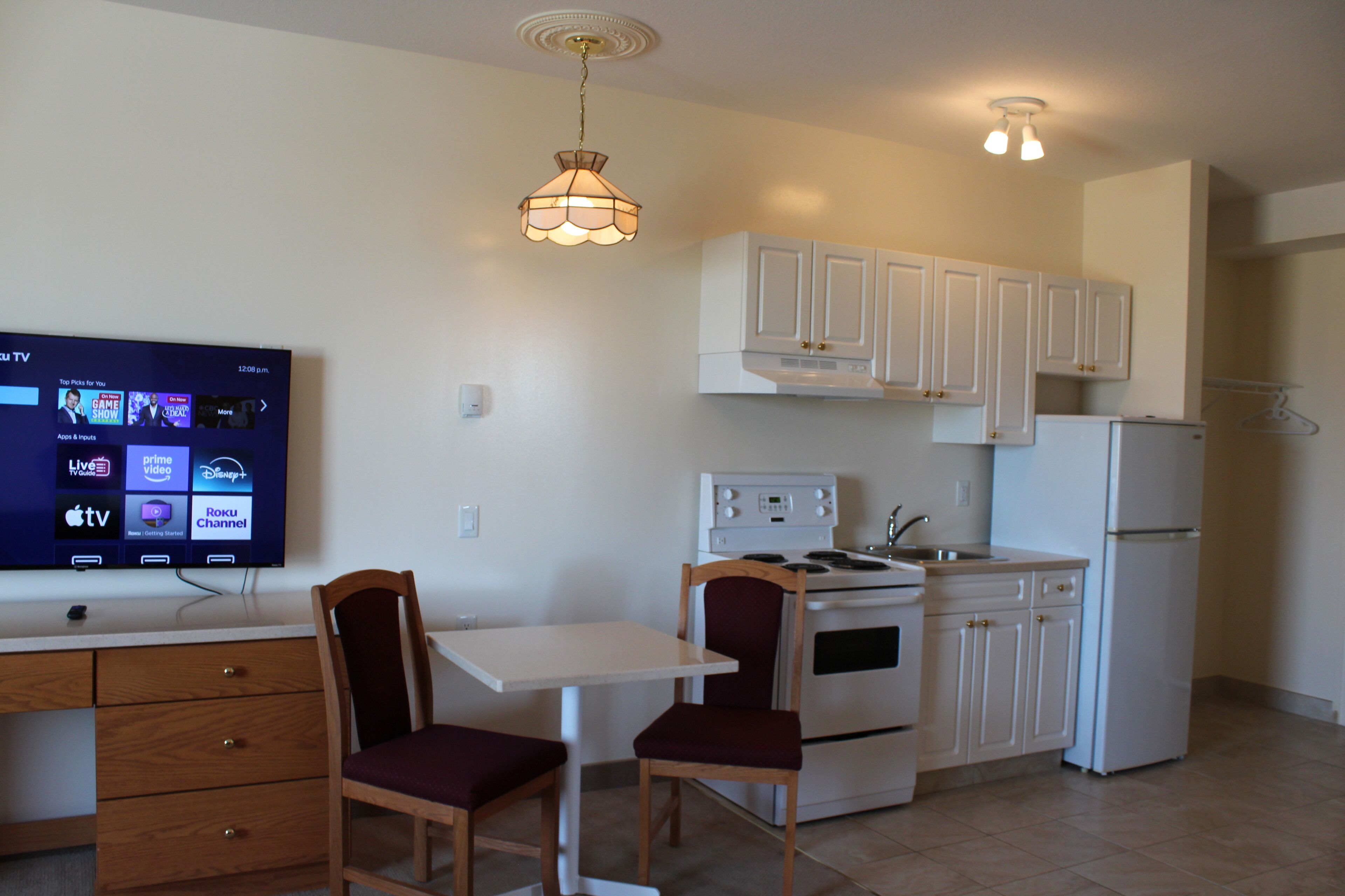 A compact kitchen with white cabinets, a stove, and a refrigerator. There's a dining table with two chairs and a TV mounted on the wall displaying various streaming services.