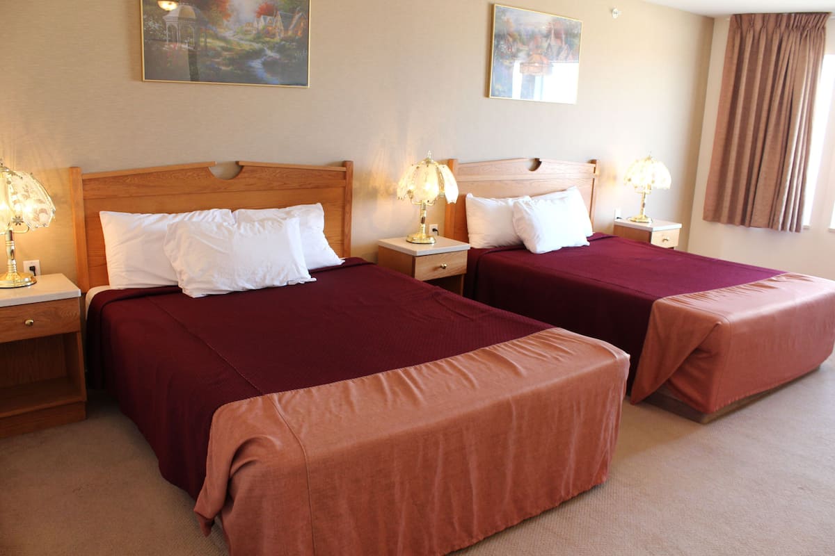A hotel room with two beds, each with a wooden headboard, white pillows, and a red bedspread. There are bedside tables with lamps and a window with curtains.