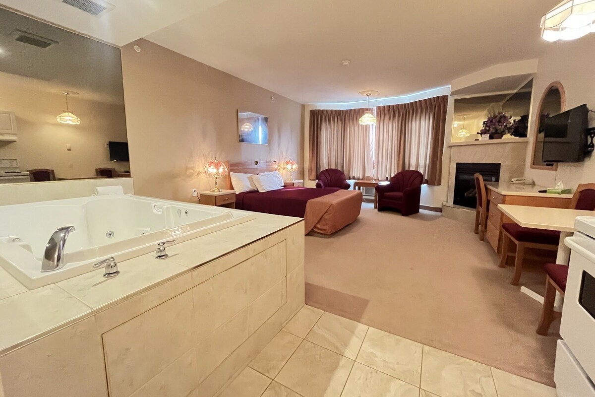 A modern hotel room with a large bathtub, a sofa, two armchairs, a dining table, and a TV.