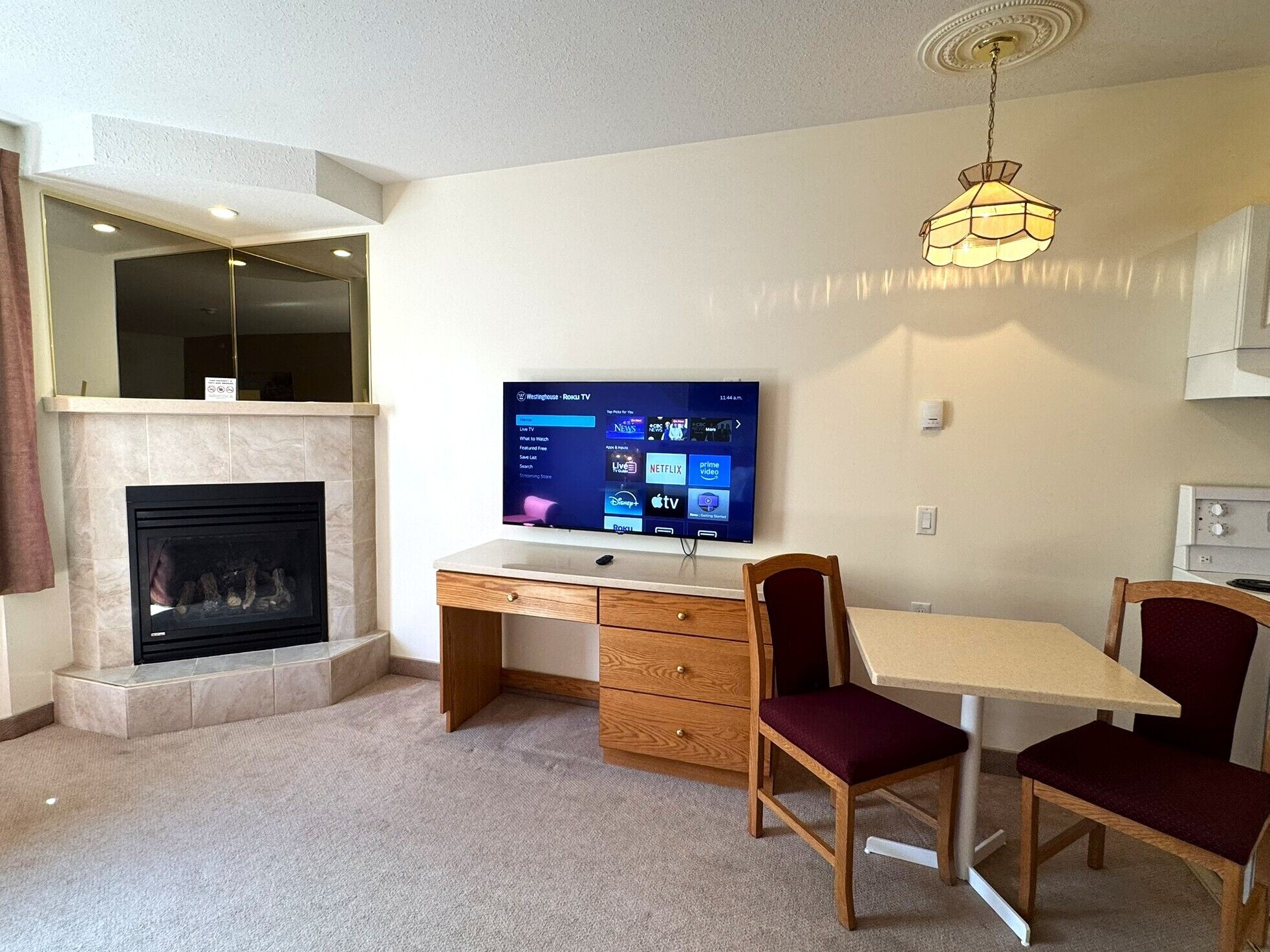 A living room with a fireplace, a TV mounted on the wall, a wooden desk, and two chairs.