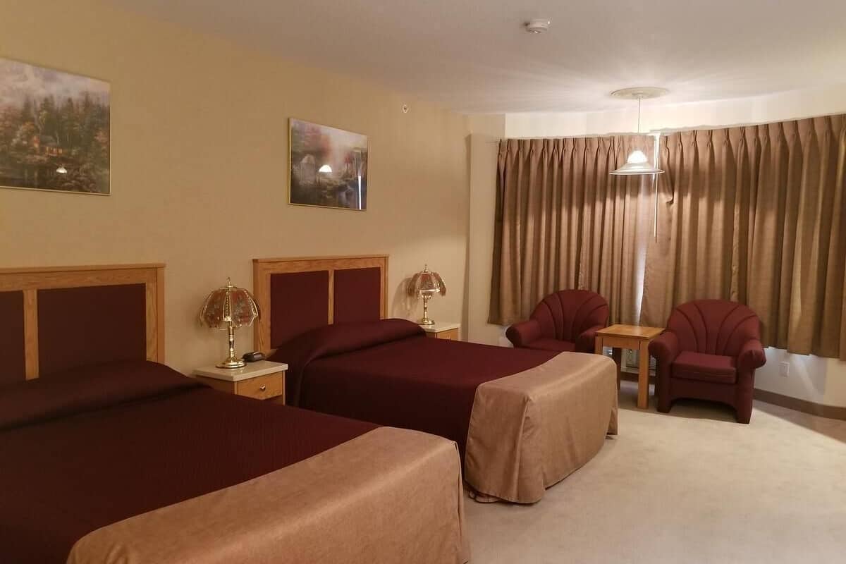 A hotel room with two beds, a chair, a table, and a large window with curtains.