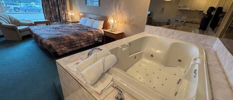 A hotel room with a large bathtub, a bed with a quilted cover, a chair, a desk, and a window with a view of parked cars outside.
