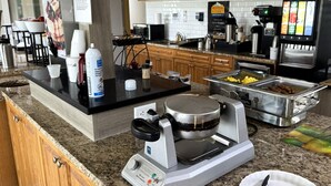 A kitchen with a coffee machine, a waffle maker, and a buffet area with food. - Western Budget Motel Whitecourt 3 (Whitecourt)
