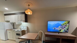 A compact kitchen with white cabinets, a stove, and a refrigerator. There's a dining table with two chairs and a television on a wooden stand. - Western Budget Whitecourt 1 & 2 (Whitecourt)