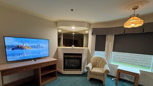 A living room with a flat-screen TV, a fireplace, a comfortable armchair, and a window with a view. - Western Budget Whitecourt 1 & 2 (Whitecourt)