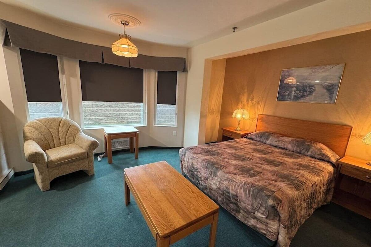A bedroom with a bed, a chair, a table, a lamp, and a painting on the wall.