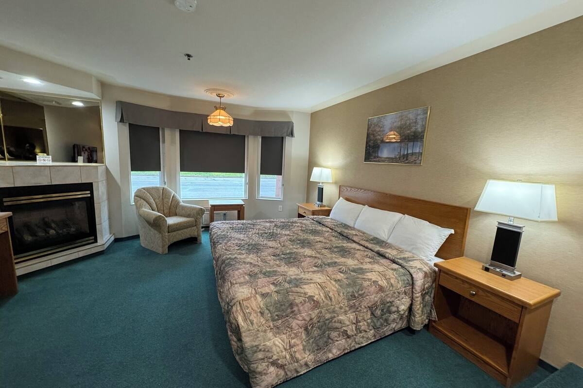 Une chambre d’hôtel avec un grand lit, une chaise, une petite table, une table de chevet, une lampe, une cheminée et un tableau au mur.
