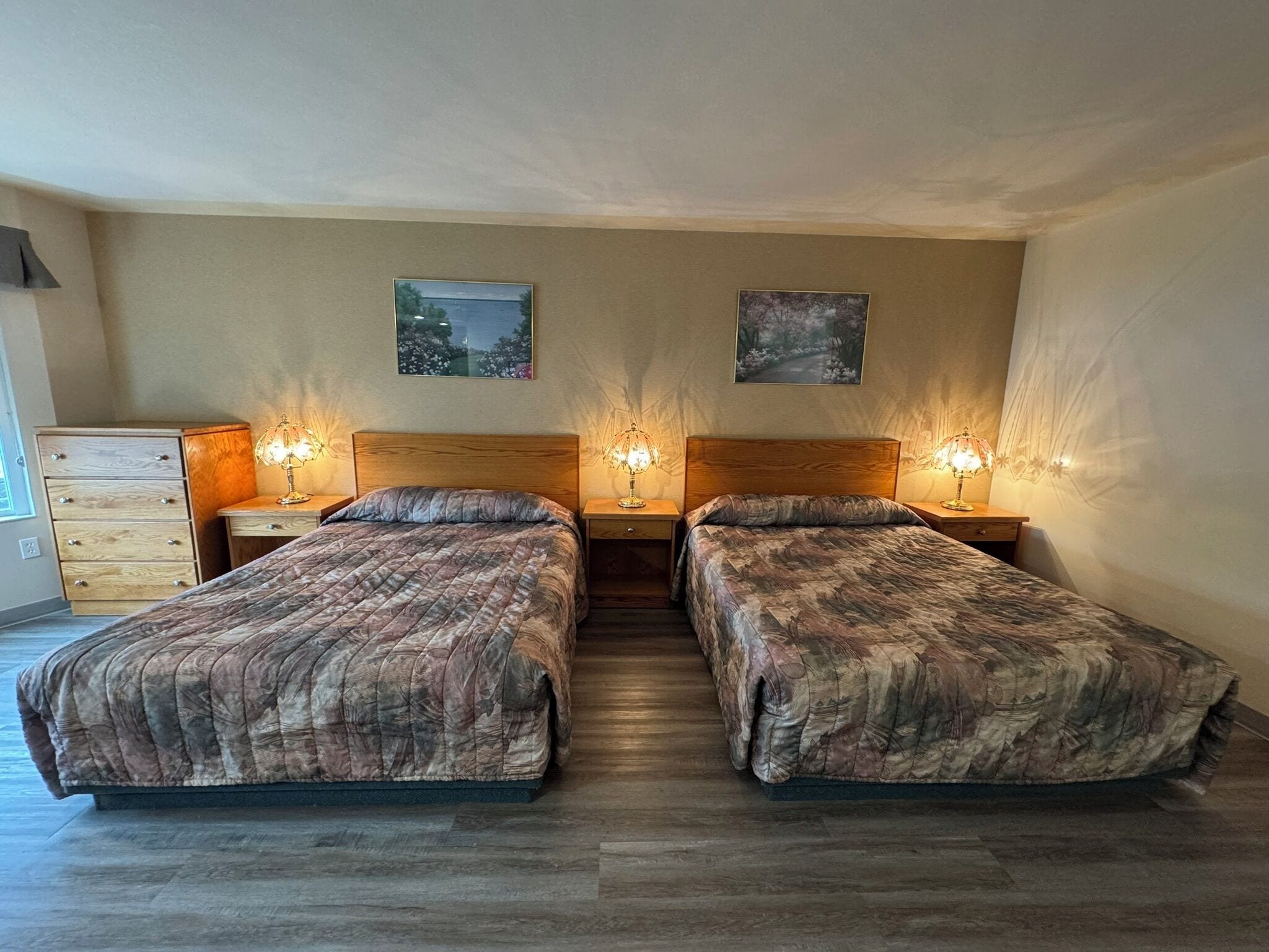 Quarto duplo com duas camas, cada uma com cabeceira e mesas de cabeceira com abajures. Há duas pinturas penduradas na parede acima das camas.