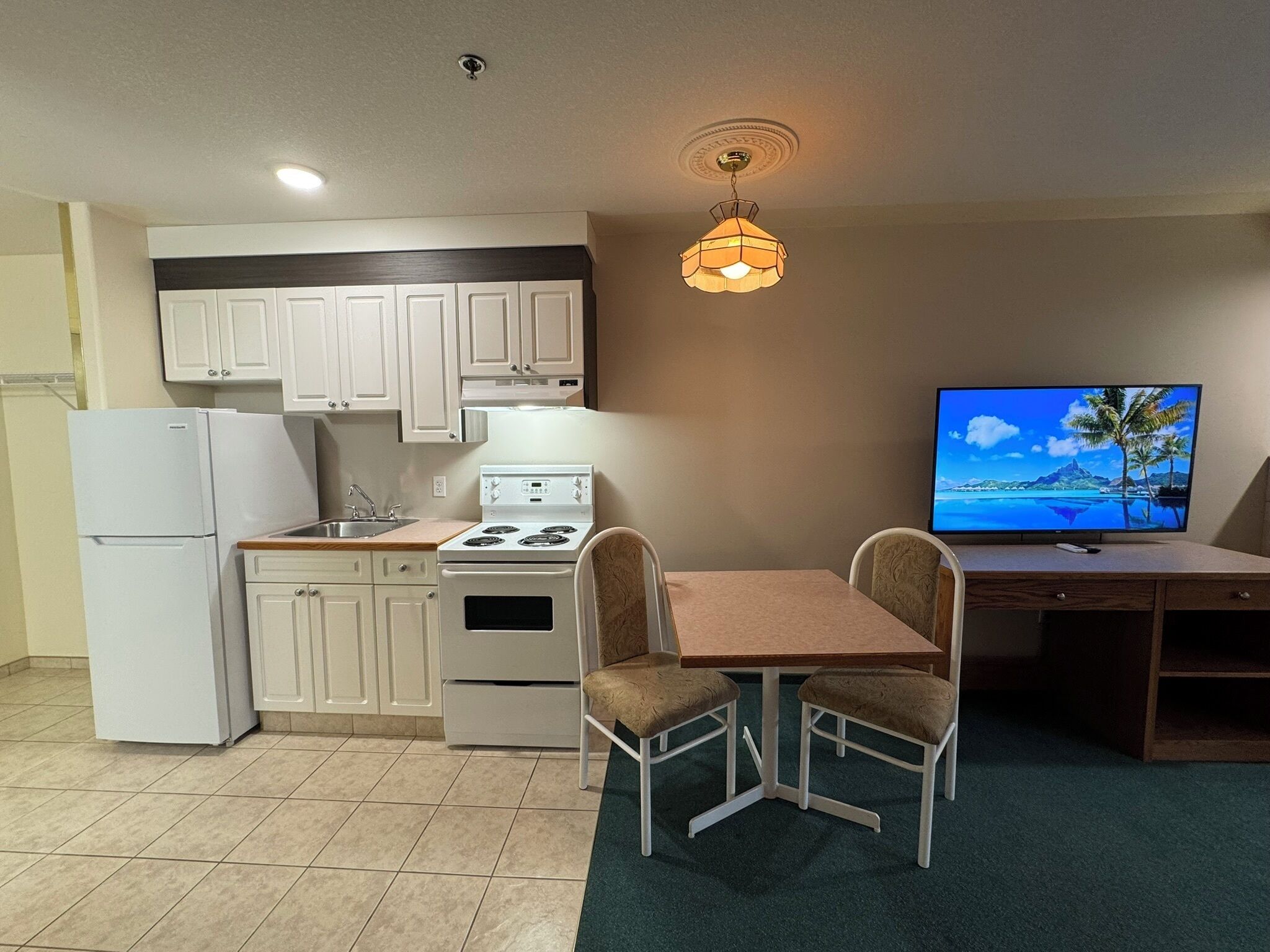 A compact kitchen with white cabinets, a stove, a sink, and a refrigerator. There is a dining table with two chairs and a television on a stand.