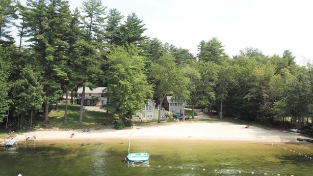 A lakeside property with a sandy beach, surrounded by dense trees. - 5 bed 4 bath in the Basin of Sebago Lake with 200 ft. of shared beach frontage (Windham)