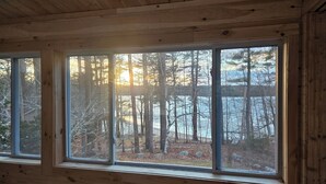 A cabin interior with a large window offering a view of a forested area. - 5 bed 4 bath in the Basin of Sebago Lake with 200 ft. of shared beach frontage (Windham)
