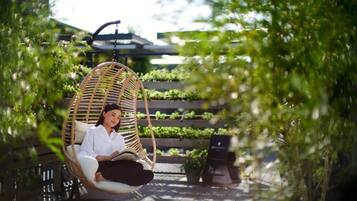 A person sitting in a wicker swing chair in an outdoor setting with a garden and a modern building in the background.