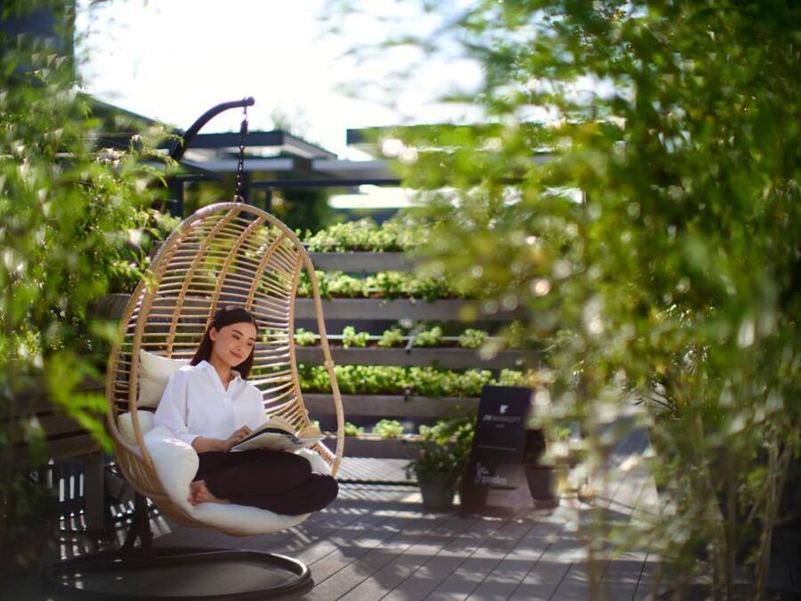 A person sitting in a wicker swing chair in an outdoor setting with a garden and a modern building in the background.