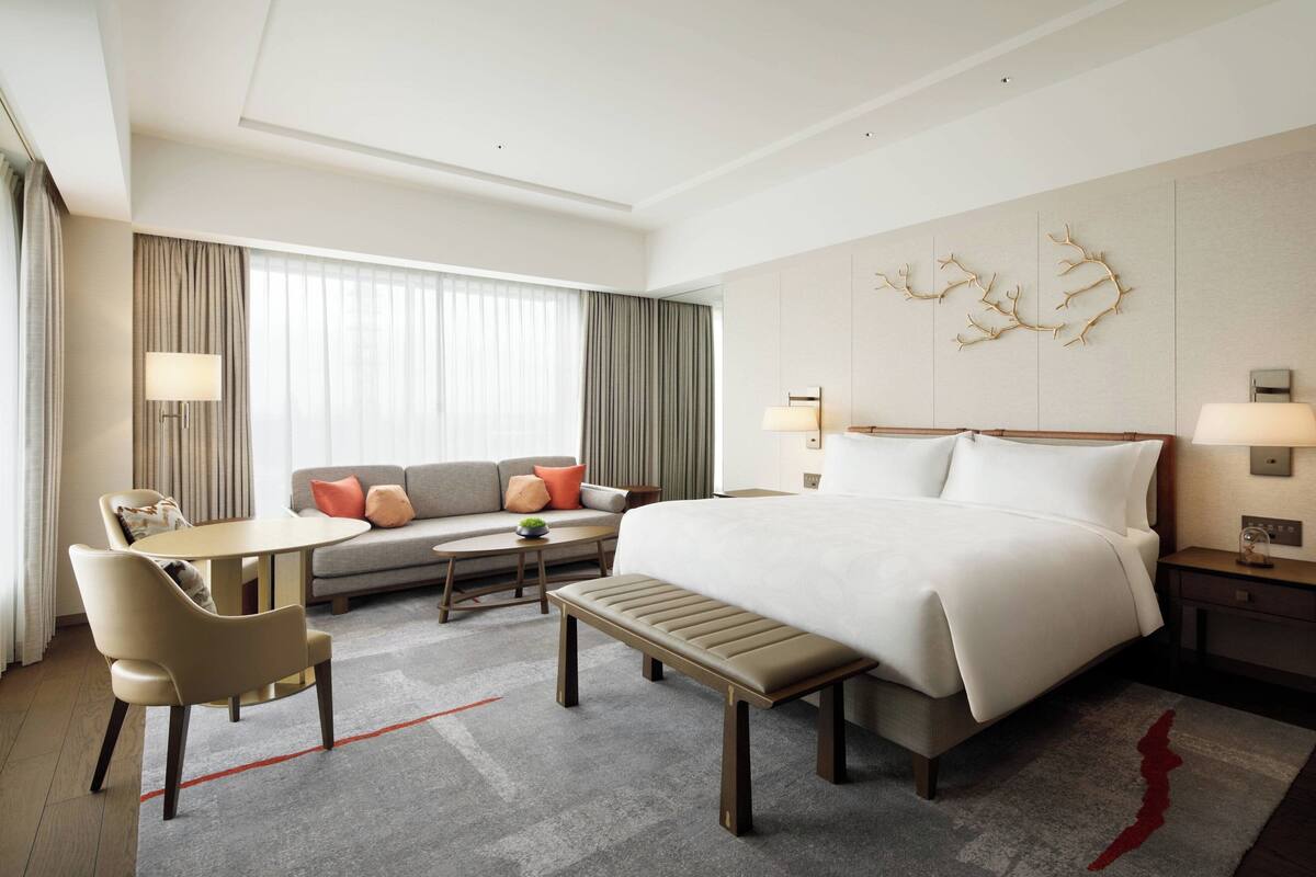 A modern hotel room with a large bed, a sofa, a desk, and a bench.