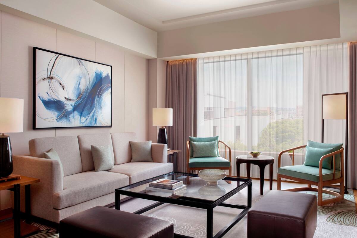 A modern hotel room with a sofa, two armchairs, a coffee table, and a large abstract painting on the wall.