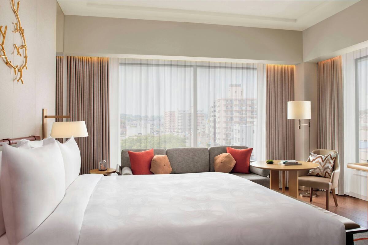 A hotel room with a large bed, a sofa, a small table, and a chair. There is a window with curtains and a view of buildings outside.