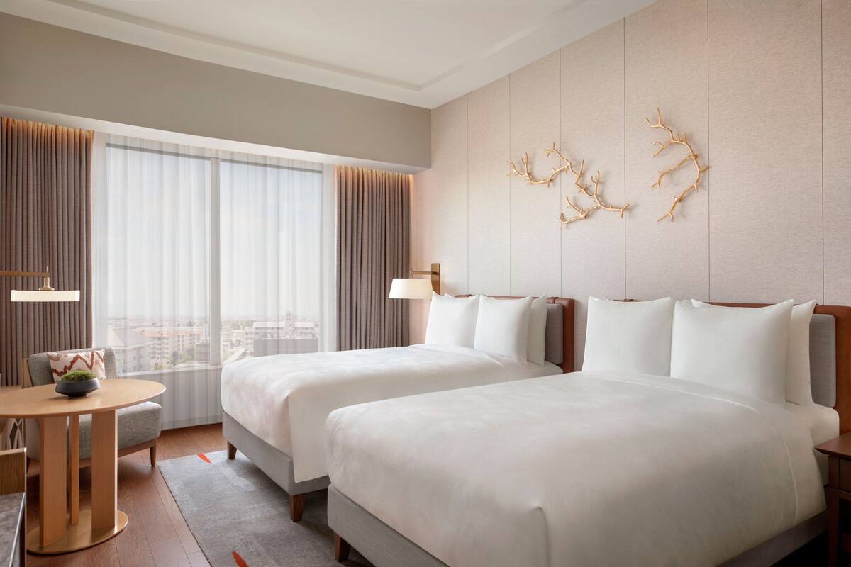A hotel room with two beds, a small table with a plant, a lamp, and a view of the city through large windows.
