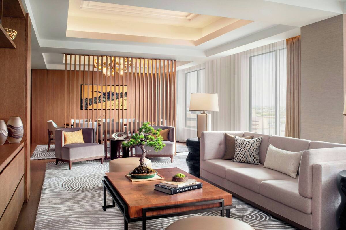 A modern hotel room with a sofa, a wooden coffee table, a bonsai tree, and a dining area with a table and chairs.