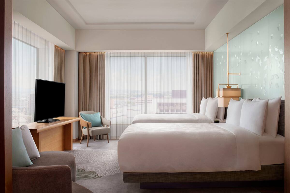 A hotel room with a large bed, a desk with a TV, a chair, and a view of the city through large windows.