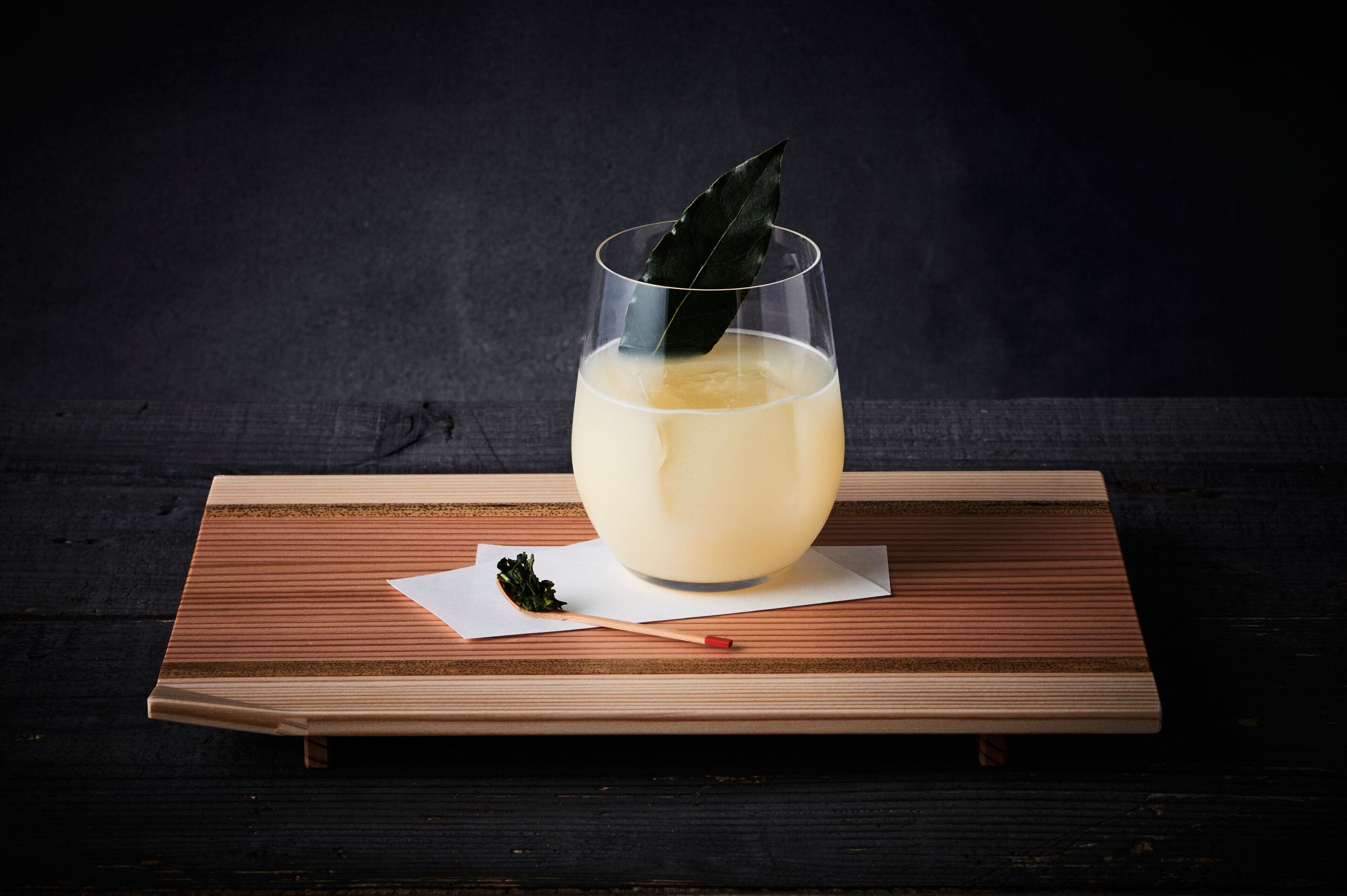 A glass of a light-colored beverage on a wooden tray with a dark background.